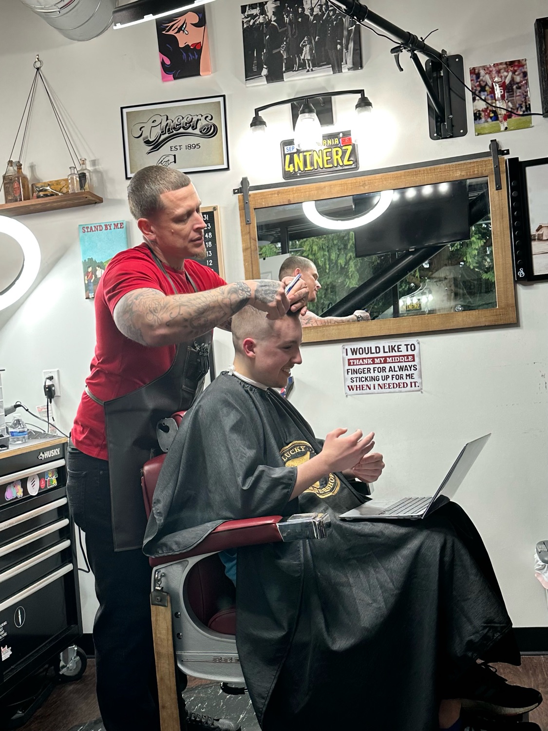 Andy in a barber's chair, mid-haircut, working on a song on his laptop while his barber cuts his hair.